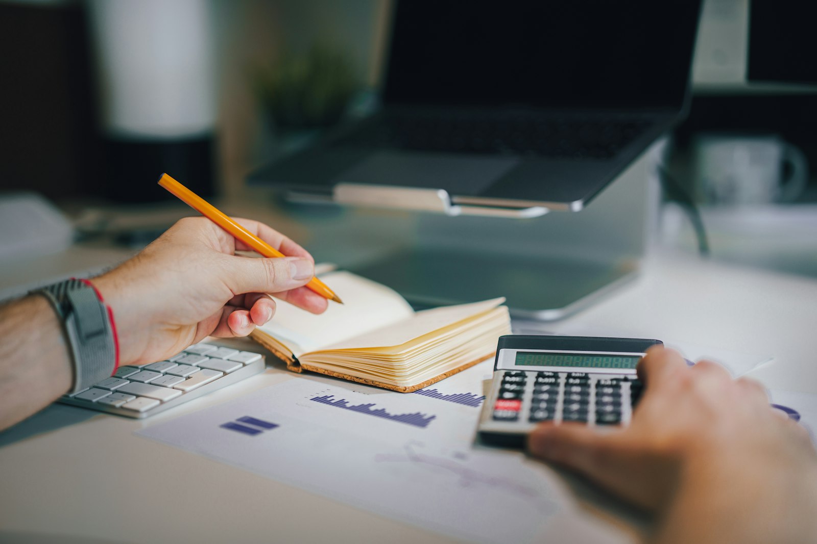 Maos de um homem usando calculadora e caderno sobre uma mesa com graficos impressos
