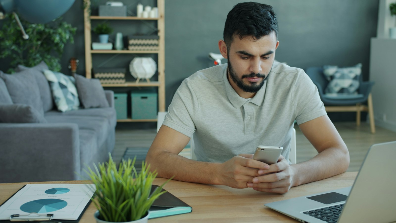 Homem conferindo o celular em uma mesa com notebook e graficos impressos