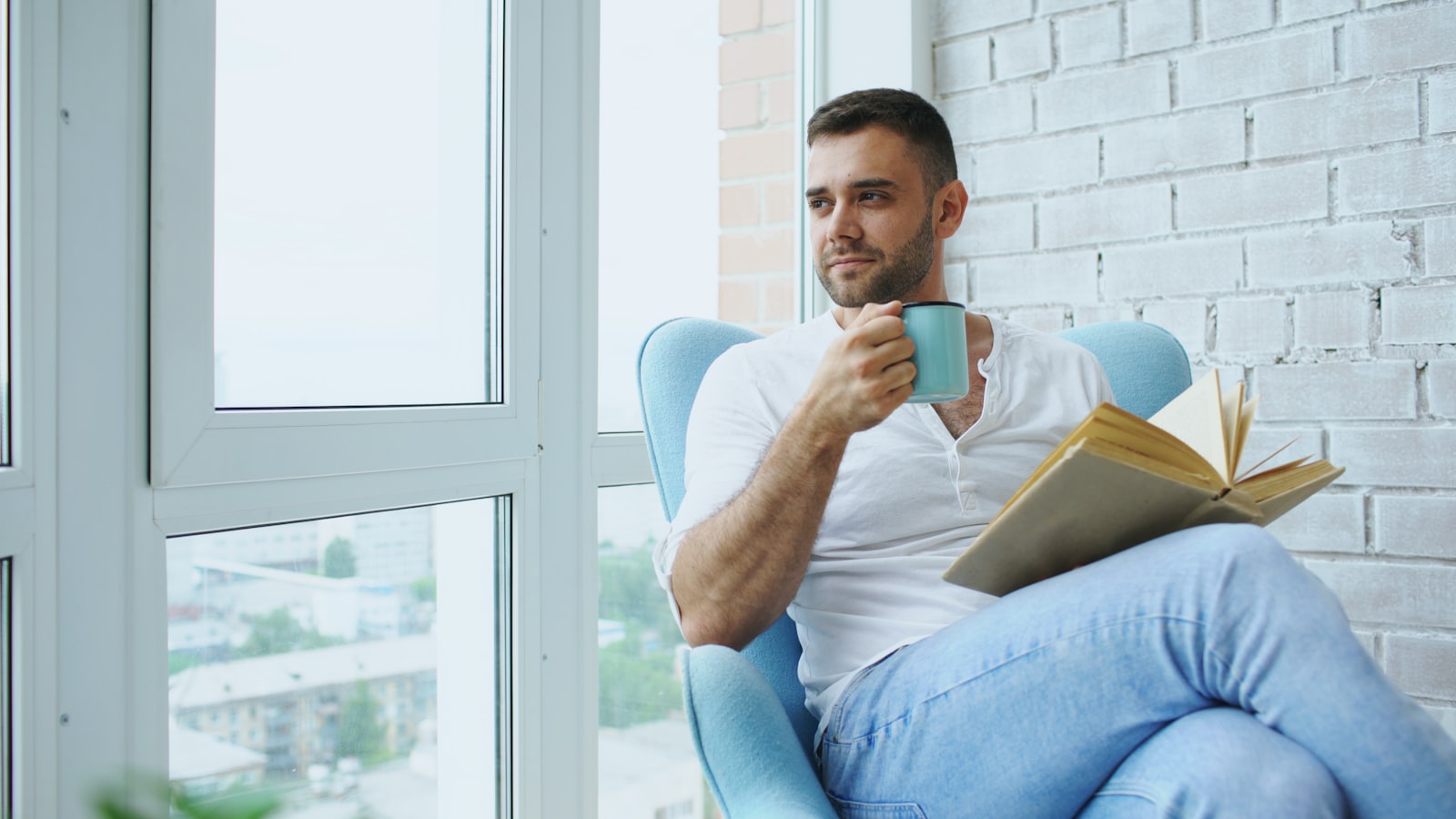 Homem sentado numa poltrona com um livro aberto e uma caneca de cafe