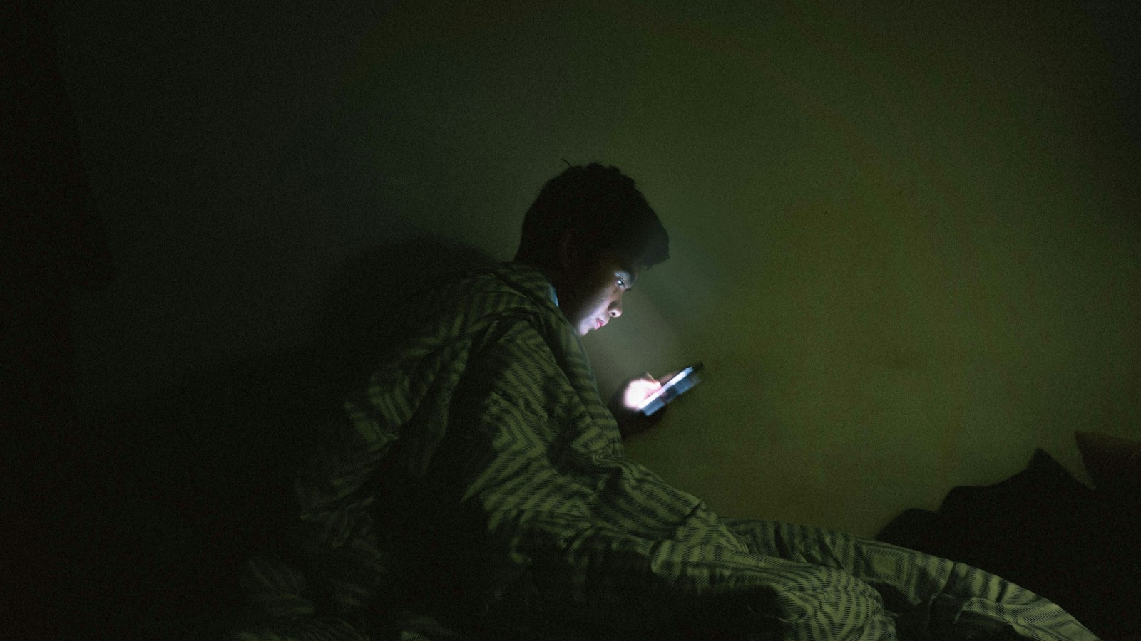 Homem deitado na cama sob o cobertor usando o celular no escuro