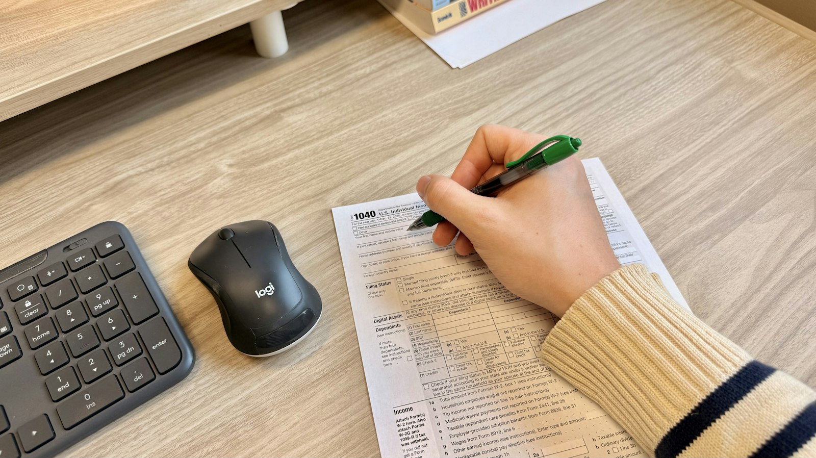 Mão preenchendo um documento impresso ao lado de teclado e mouse em uma mesa de trabalho
