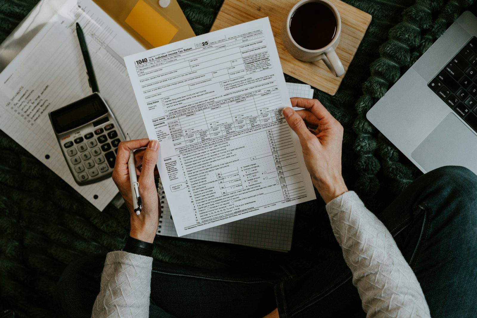 Mãos segurando formulários de imposto ao lado de uma calculadora e um notebook sobre a mesa