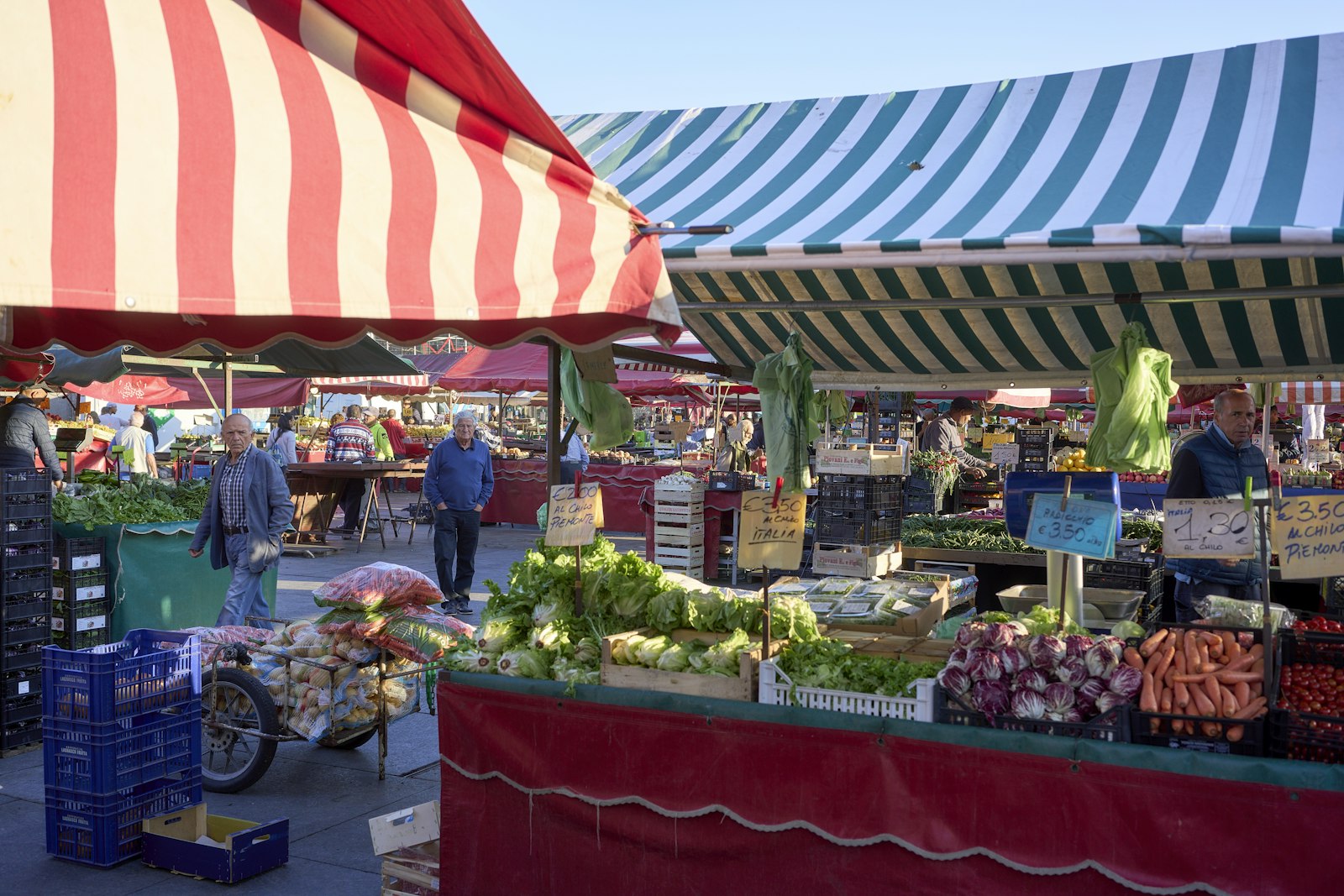 Barracas de feira livre com verduras e legumes expostos em caixas sob toldos listrados