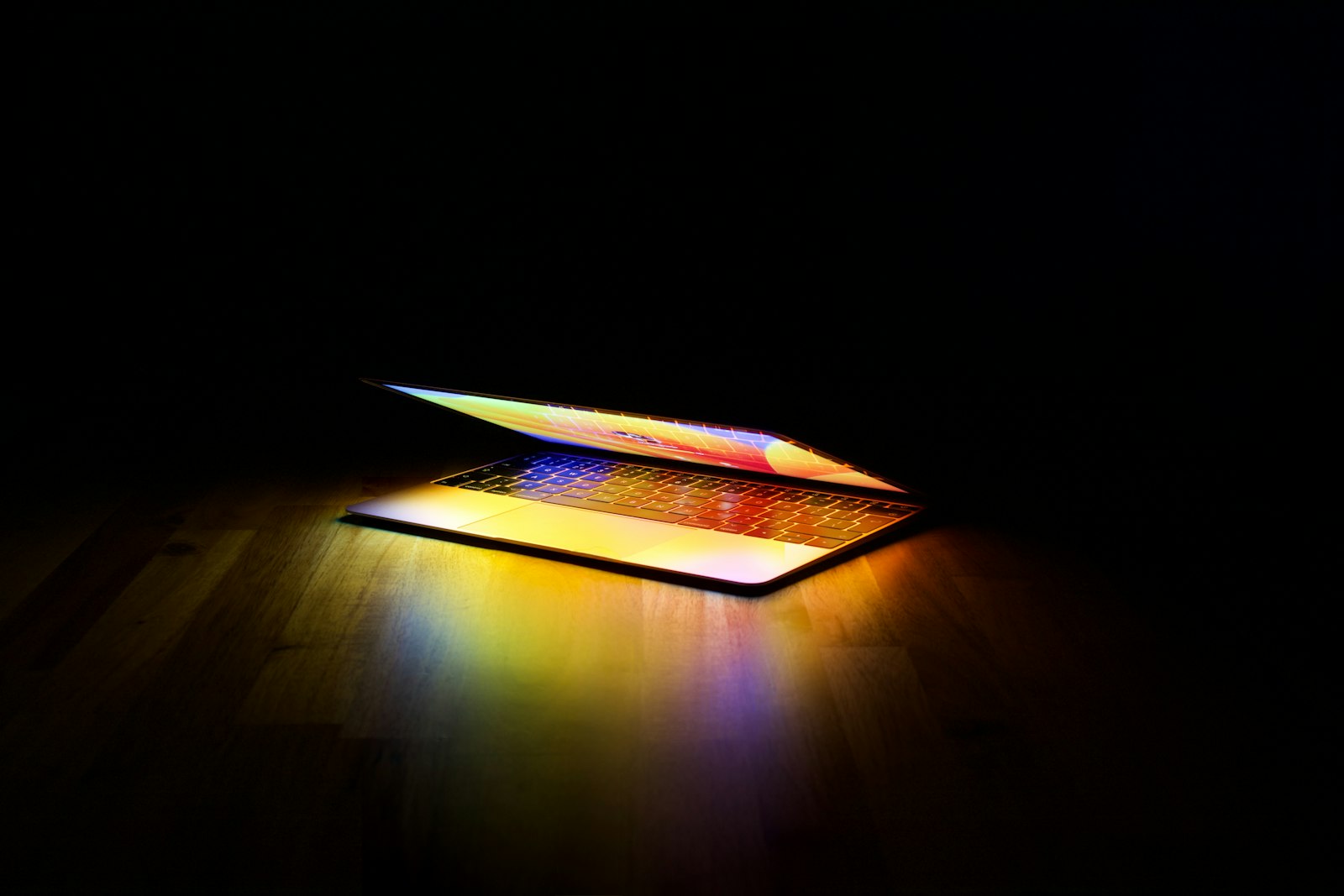 MacBook aberto iluminando uma mesa de madeira num quarto escuro